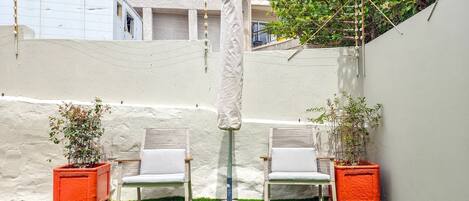 A small outdoor seating area with two white chairs and a table, surrounded by potted plants and a white wall.
