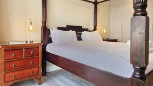 Een hemelbed met houten frame, een nachtkastje met lamp en een houten dressoir met lades.