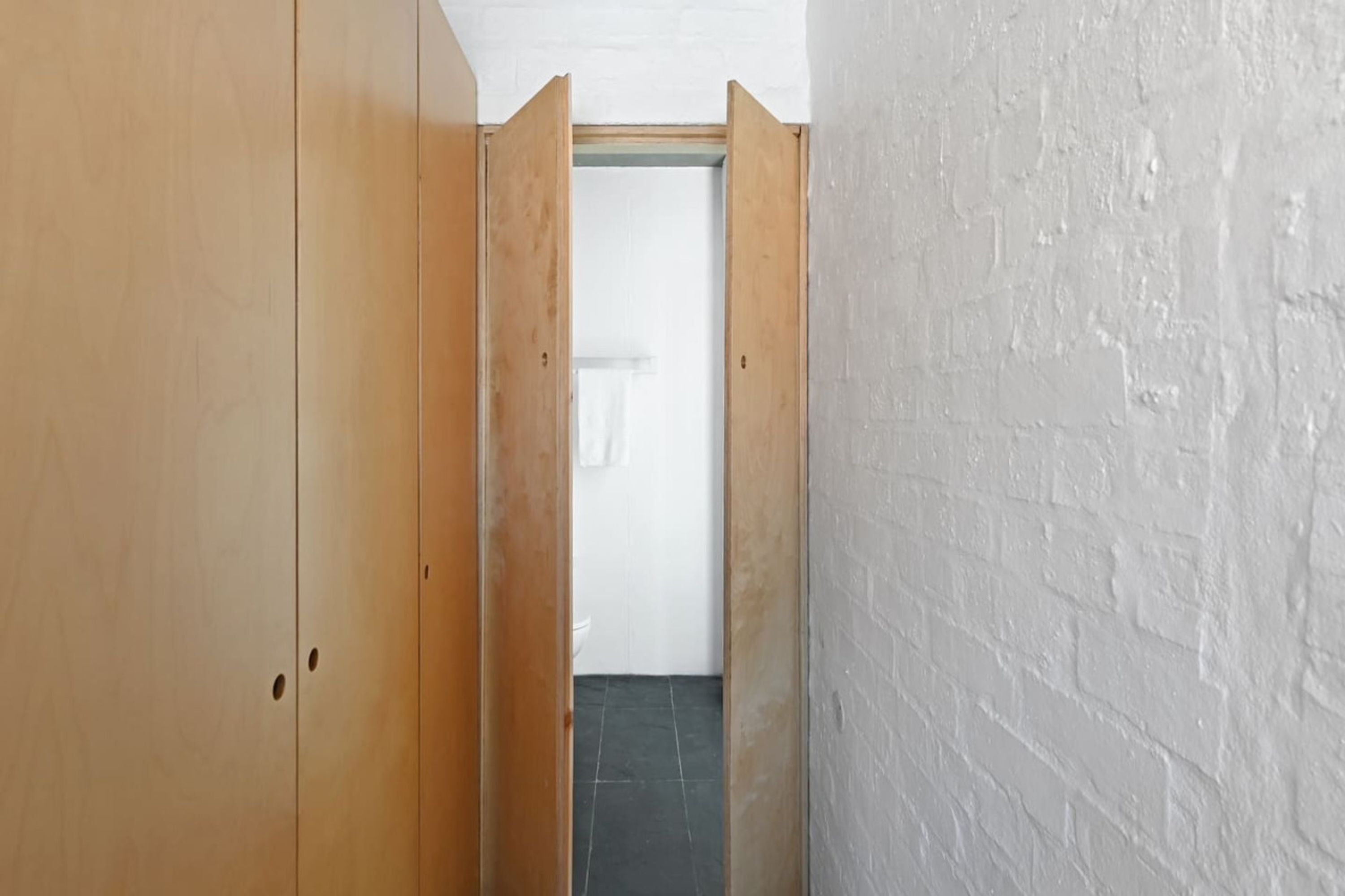 Um corredor estreito, com uma porta de madeira à esquerda e uma parede de tijolos brancos à direita.