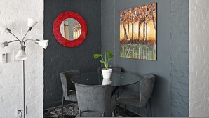 Un'area pranzo con un tavolo rotondo, sedie grigie, una pianta in vaso, uno specchio con cornice rossa e un quadro colorato appeso al muro.