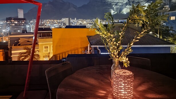 Eine Dachterrasse mit Tisch, Stühlen und einem Blumenstrauß. Eine rote Markise ist links zu sehen. Im Hintergrund erstreckt sich eine Stadtlandschaft mit Gebäuden und Bergen.