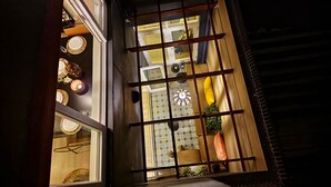 Blick durch ein Fenster mit Holzrahmen und Metallgitter, das in einen Raum mit gemustertem Wandpapier, einer Hängeleuchte und einem Blumentopf führt.