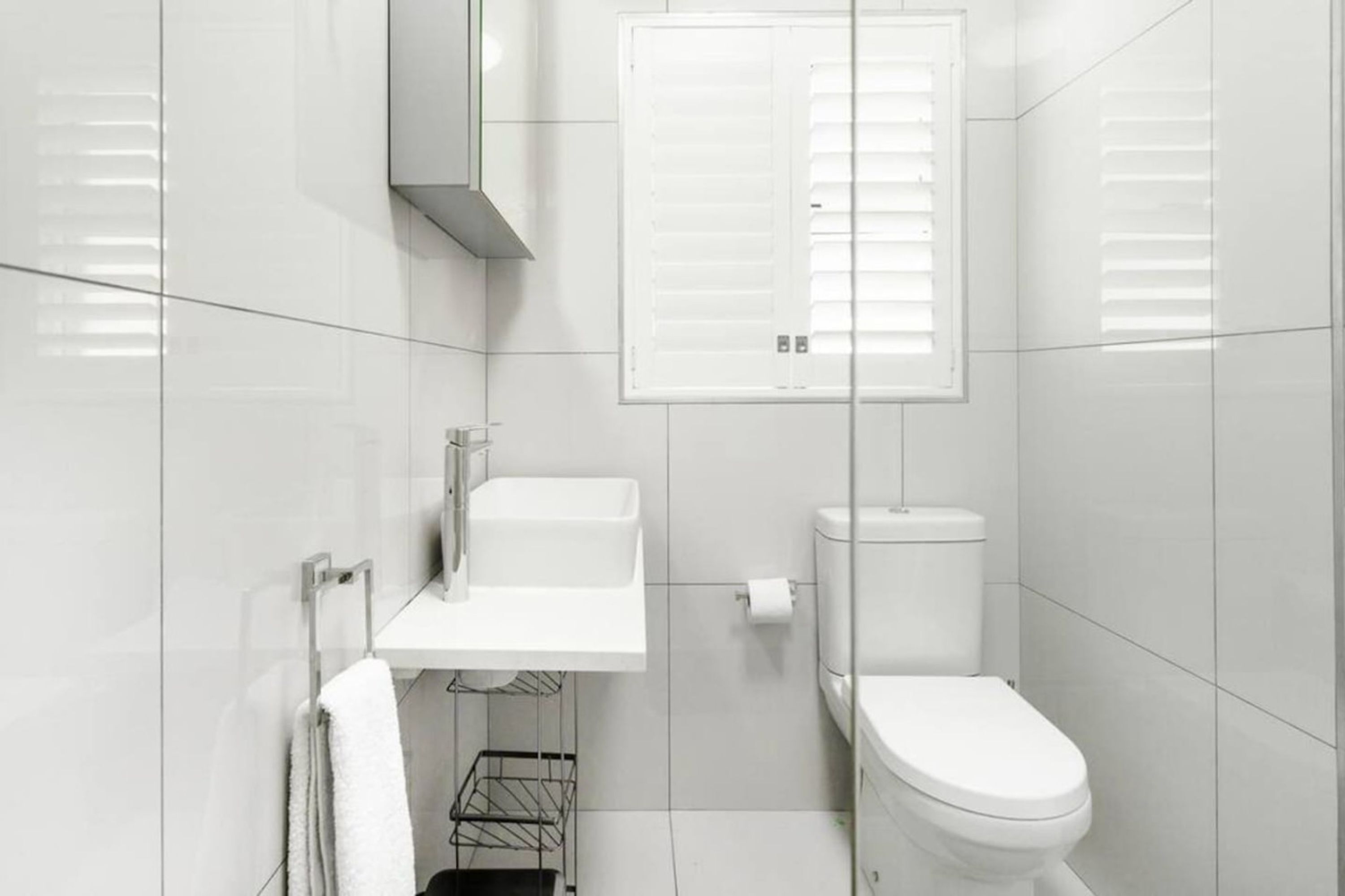 Ein modernes Badezimmer mit weißen Fliesen, einer Toilette, einem Waschbecken mit Spiegel darüber und einem Fenster mit Jalousien.