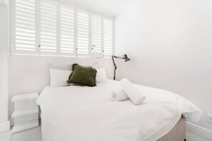 Een slaapkamer met een bed, witte beddengoed, een groen kussen, een bedlampje en een raam met jaloezieën.
