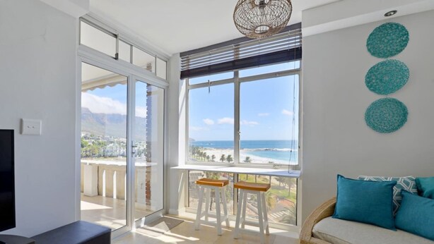 A spacious balcony with a view of the beach and mountains, a sofa with cushions, and a television. - Top-floor 2-bed Beachfront Sunny Camps Bay Views (Cape Town)
