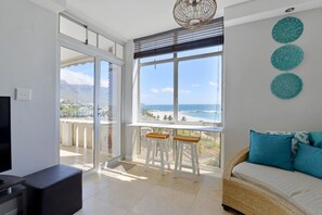 Un balcon spacieux offrant une vue sur la plage et les montagnes, un canapé avec des coussins et un téléviseur.