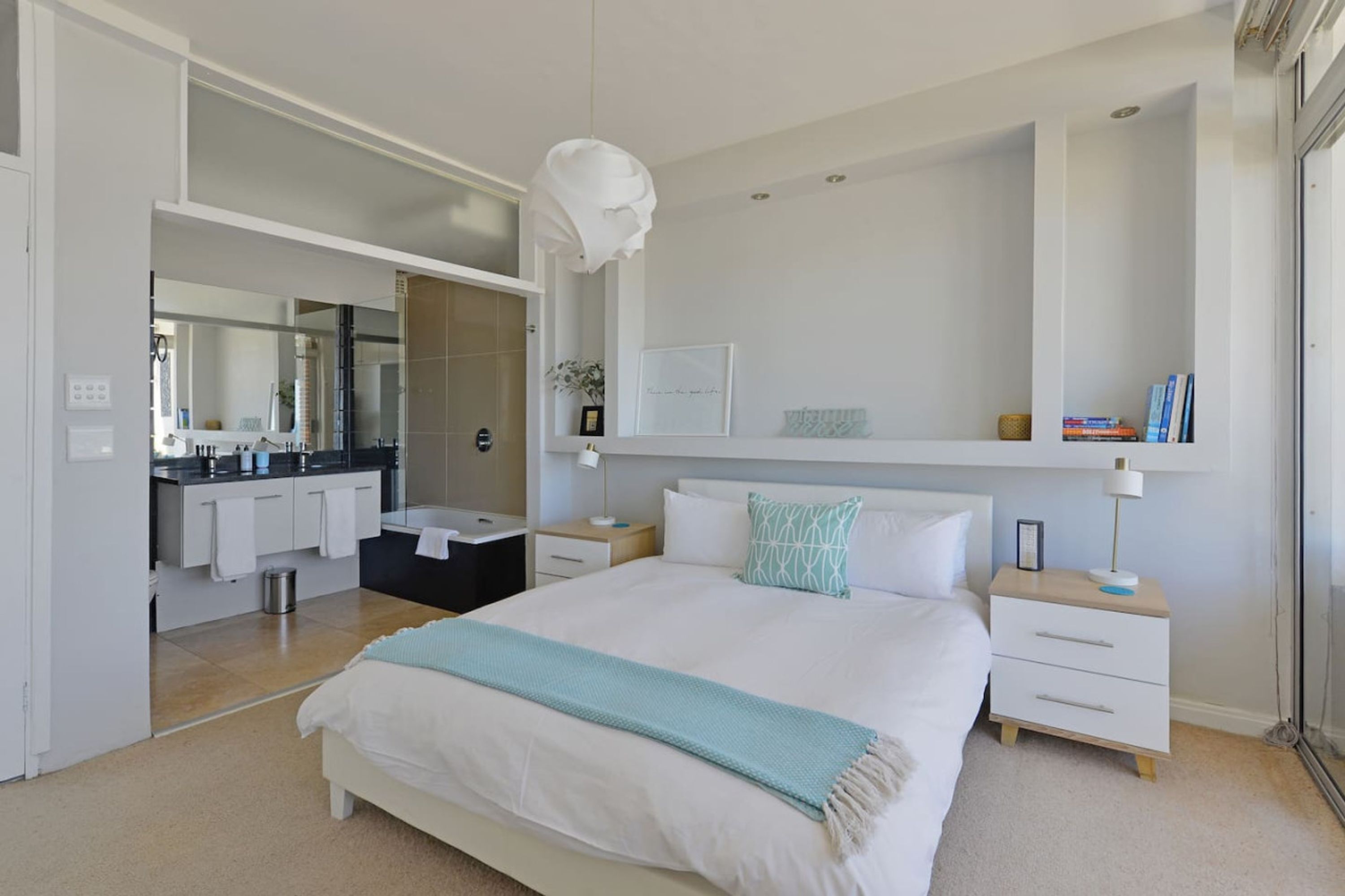 A modern bedroom with a large bed, bedside tables, a nightstand, a mirror, and a built-in shelf with books.