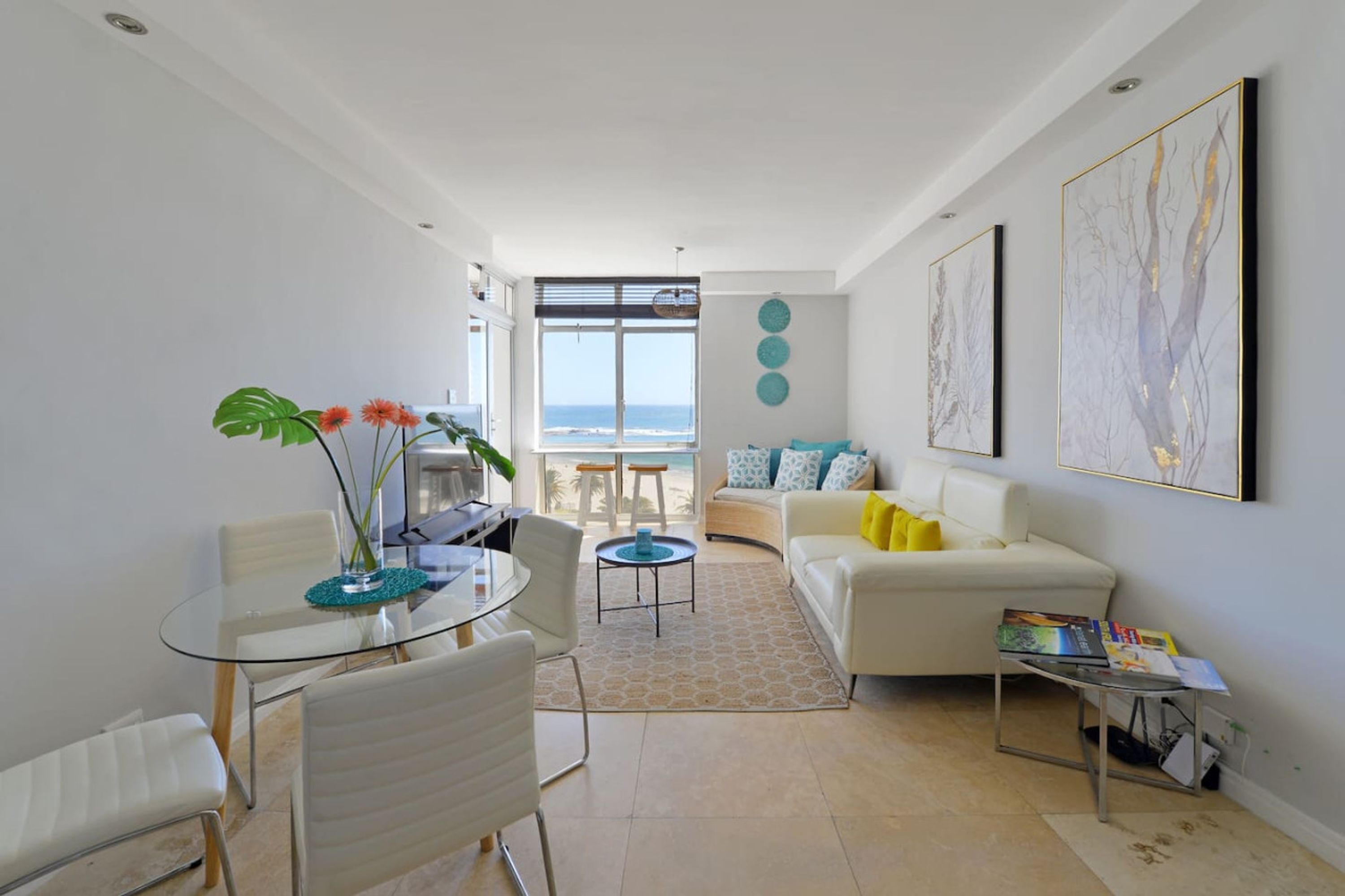 A modern living room with a glass dining table, white sofas, and a view of the ocean.