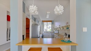 A modern kitchen with a wooden countertop, two white bar stools, hanging white pendant lights, and a view of the outside through a window. - Top-floor 2-bed Beachfront Sunny Camps Bay Views (Cape Town)
