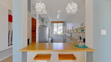 A modern kitchen with a wooden countertop, two white bar stools, hanging white pendant lights, and a view of the outside through a window.