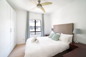 A hotel room with a bed, a ceiling fan, a window with curtains, and a nightstand with a lamp. - 5th Floor - City Center 3bed Central and Spacious (Cape Town)