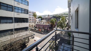 Un immeuble à plusieurs étages doté d’un balcon donnant sur une rue où sont garées des voitures, avec en arrière-plan un mélange de bâtiments commerciaux et résidentiels.