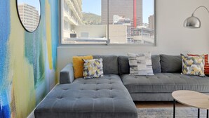 A modern living room with a grey sectional sofa, a round mirror, and a view of city buildings through large windows. - Refined City Living Sunny Pool Gym Facilities (Cape Town)