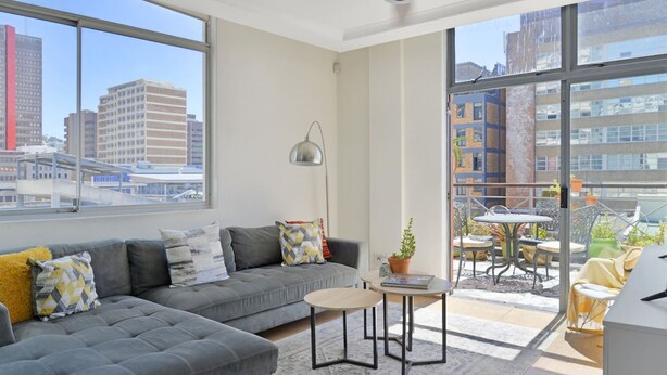 A modern living room with a grey sectional sofa, a small round table, and a balcony with outdoor seating. - Refined City Living Sunny Pool Gym Facilities (Cape Town)