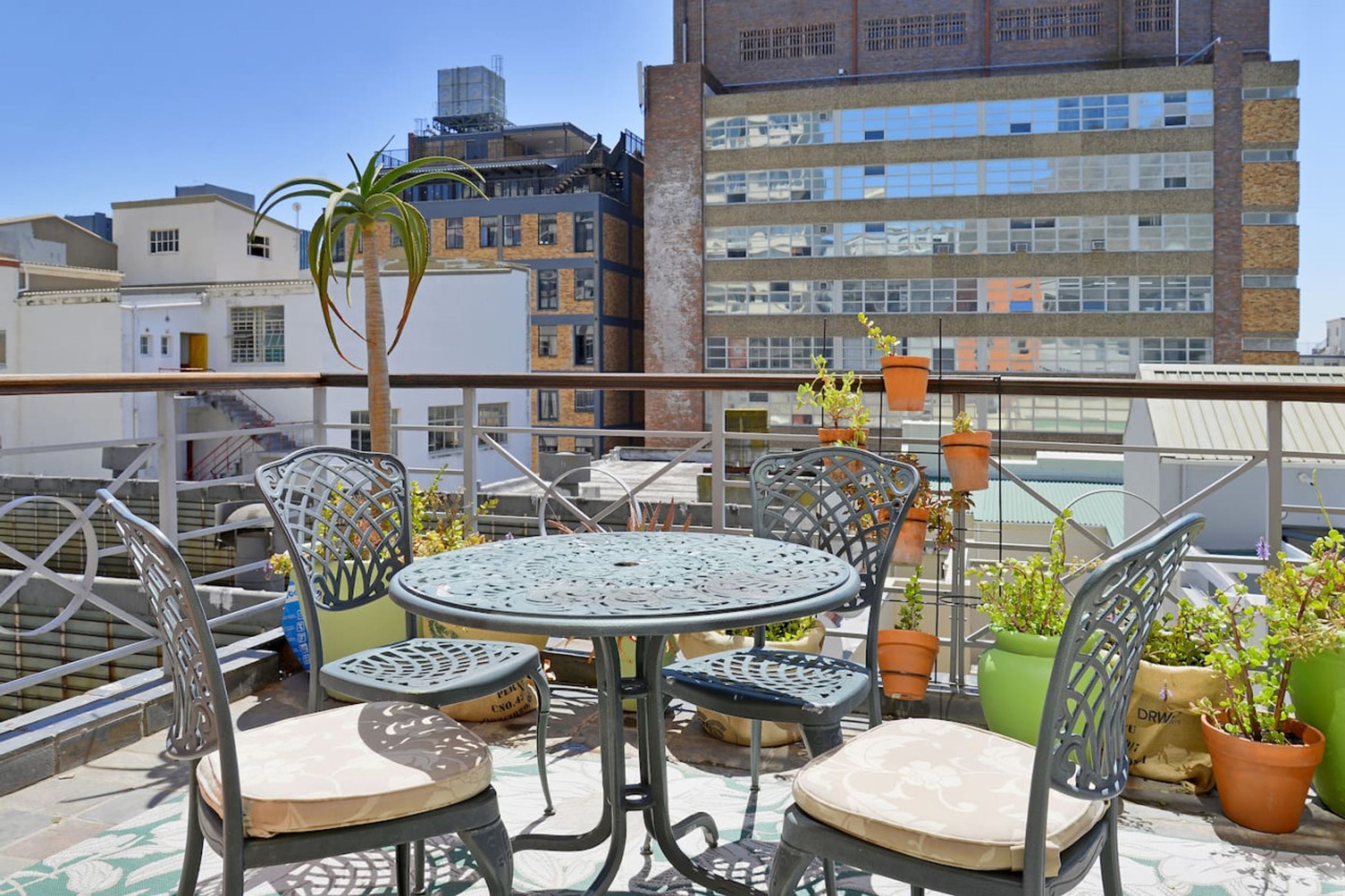 A rooftop terrace with a round table and chairs, surrounded by potted plants and a view of a cityscape.