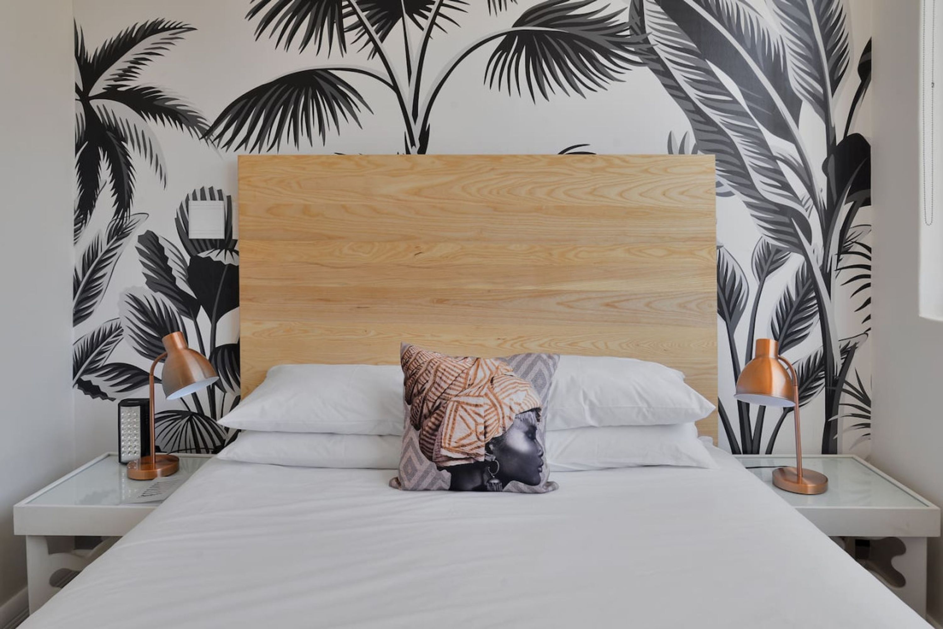 Un dormitorio con cabecera de madera, ropa de cama blanca, un cojín estampado y dos lámparas de noche.