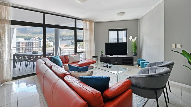 A modern living room with a red leather sofa, a glass coffee table, and a grey armchair. A flat-screen TV is mounted on the wall, and there is a balcony with a dining table and chairs. - Modern 3-bed Apt Designer Touch Mountain Views (Cape Town)