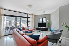 A modern living room with a red leather sofa, a glass coffee table, and a grey armchair. A flat-screen TV is mounted on the wall, and there is a balcony with a dining table and chairs. - Modern 3-bed Apt Designer Touch Mountain Views (Cape Town)