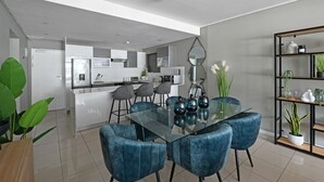 A modern kitchen with white cabinets, a central island, and a dining area with a glass table and blue chairs. - Modern 3-bed Apt Designer Touch Mountain Views (Cape Town)