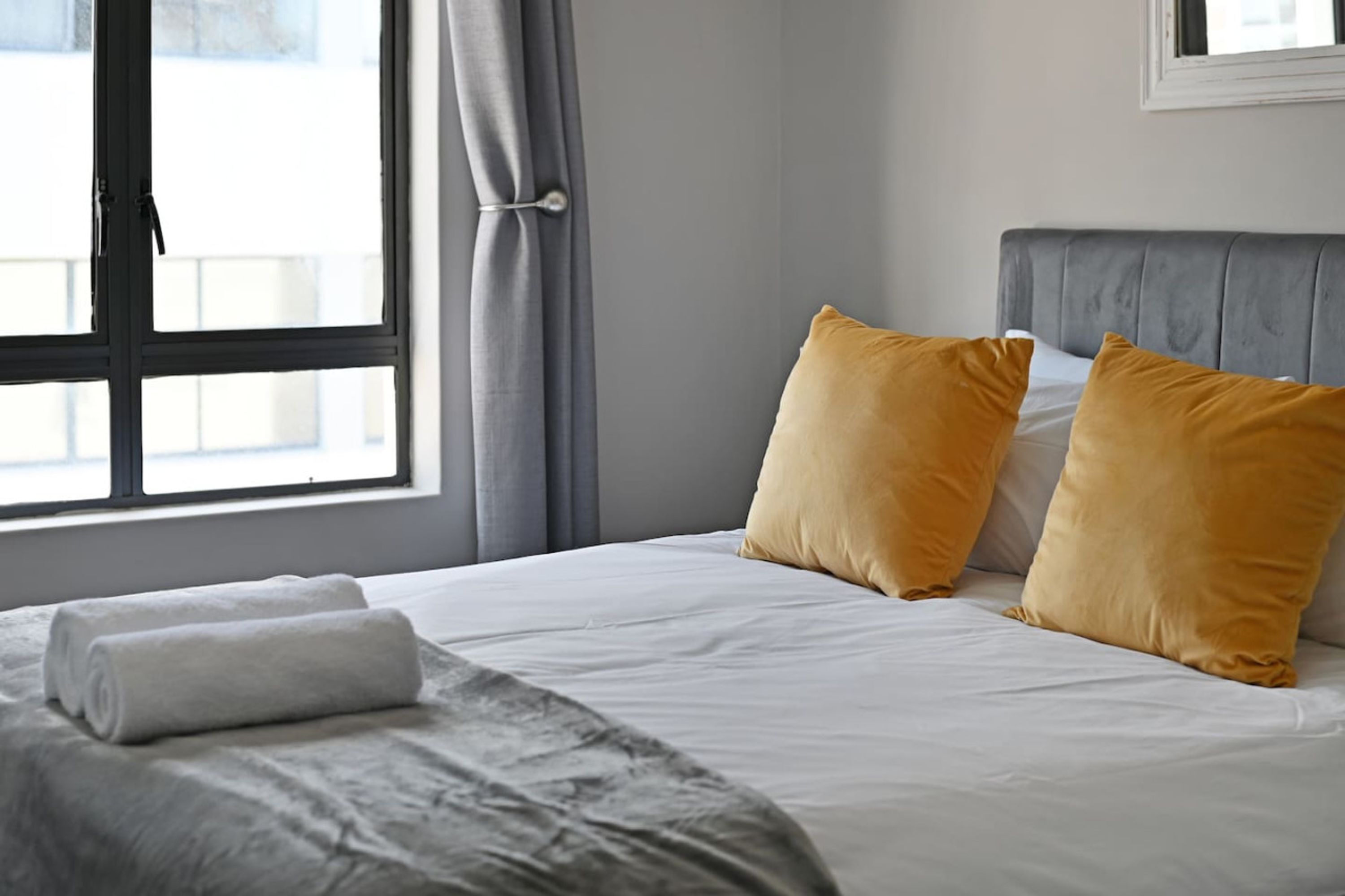 Una cama bien hecha con ropa de cama blanca y dos almohadas color mostaza, un cabecero gris y una ventana con cortinas grises.