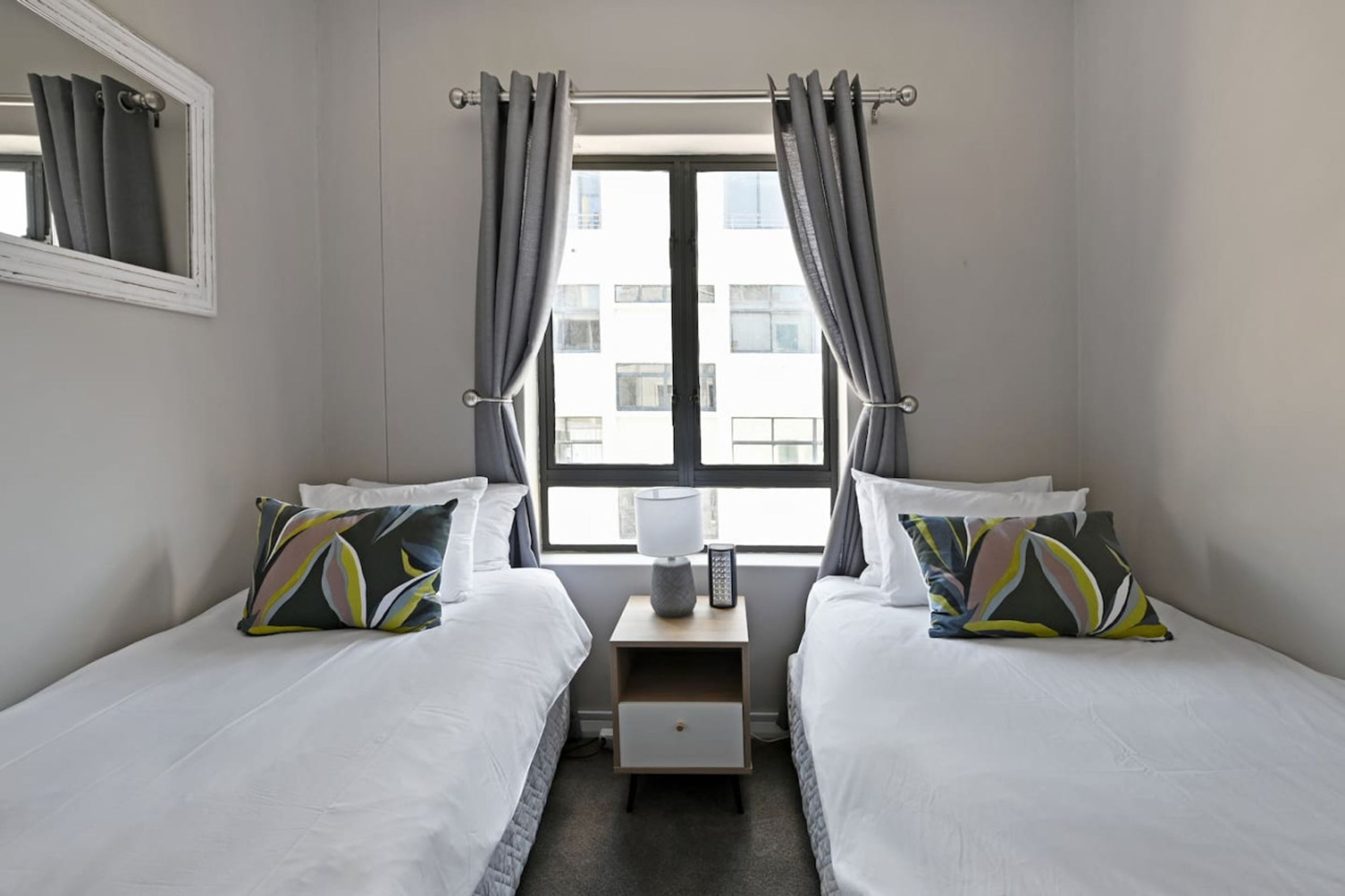 A hotel room with two beds, a nightstand with a lamp, and a window with curtains.
