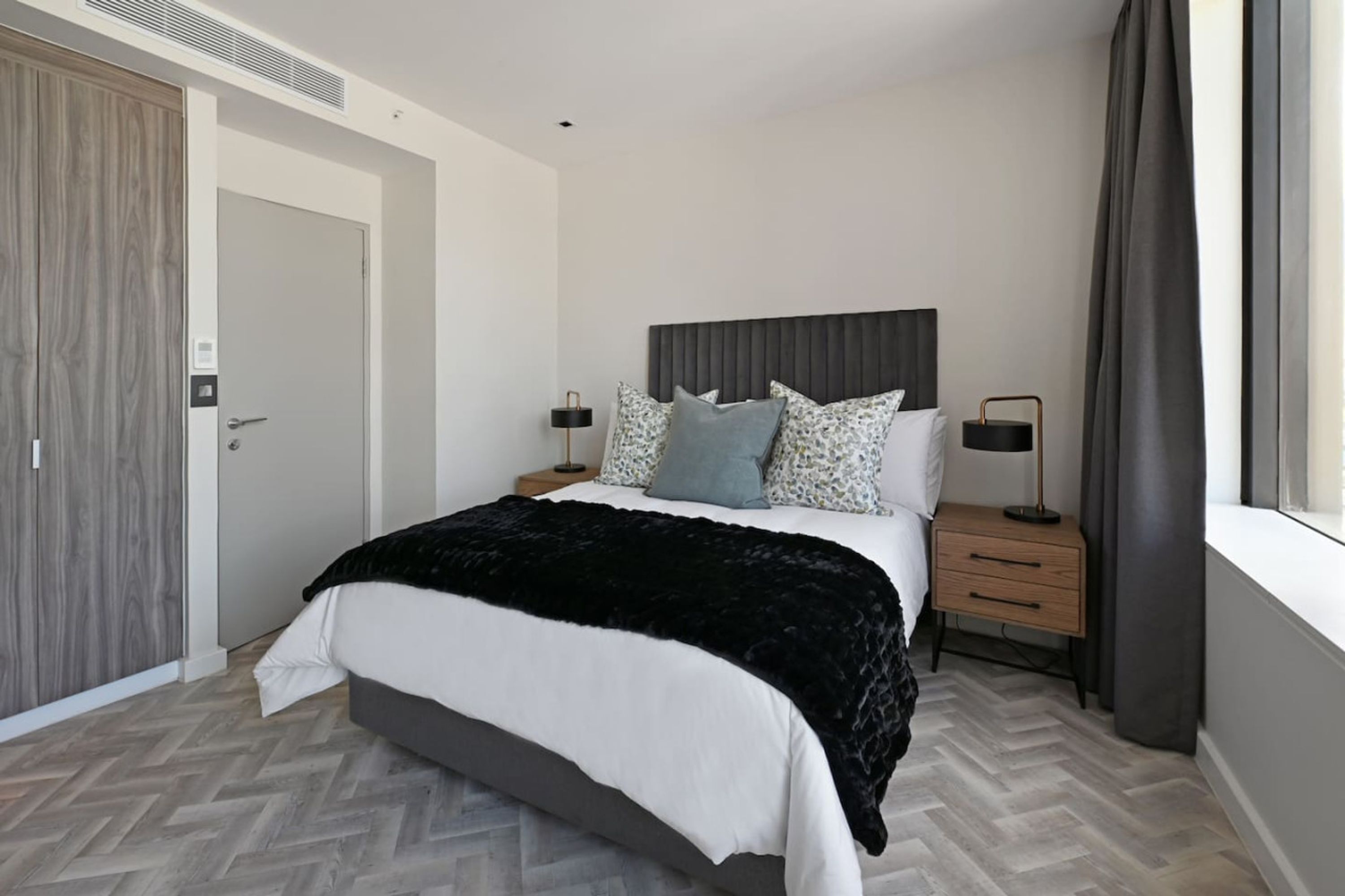 A modern bedroom with a large bed, a wooden wardrobe, a bedside table with a lamp, and a window with curtains.