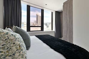 Una habitación de hotel moderna con una cama grande, cortinas oscuras y vistas a la ciudad a través de un ventanal amplio.