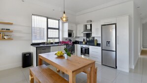 A modern kitchen with white cabinetry, stainless steel appliances, a wooden dining table, and a black trash bin. - Modern 2-bed Green Point Haven Stylish Central (Cape Town)