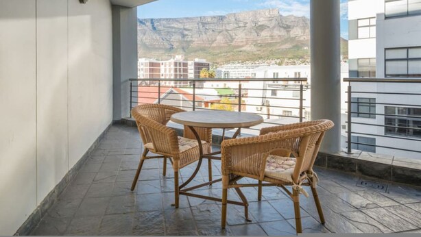 A balcony with wicker chairs and a table, overlooking a cityscape with buildings and a mountain in the background. - 6th Floor - 3bed City Center Spacious Secure (Cape Town)