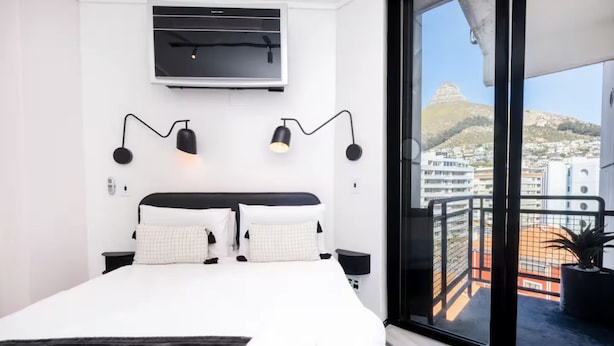 A modern bedroom with a large bed, a mounted TV, two wall-mounted lamps, and a balcony with a view of buildings and a hill. - The Flamingo One Bed Sea Point Promenade (Cape Town)