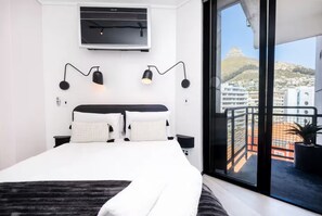 Una camera da letto moderna con un letto grande, una TV a parete, due lampade a parete e un balcone con vista su edifici e una collina.