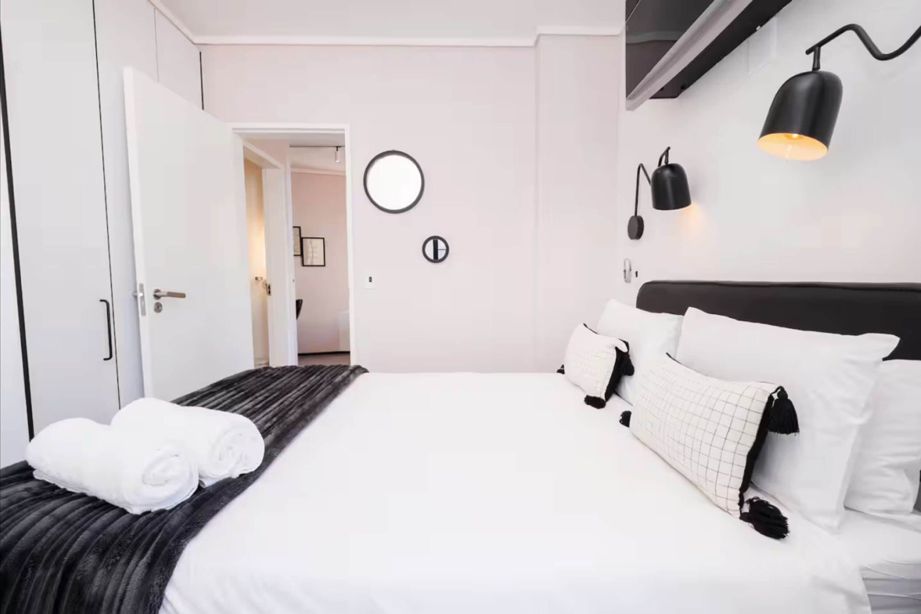 A modern hotel room with a neatly made bed, a round mirror, and a wall-mounted lamp.