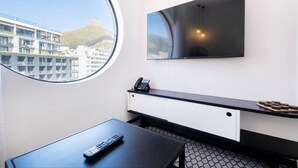 A hotel room with a large window, a flat-screen TV mounted on the wall, a desk with a telephone, and a remote control on the table. - The Flamingo One Bed Sea Point Promenade (Cape Town)