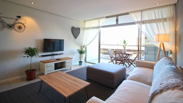 A modern living room with a sofa, a coffee table, a TV, and a balcony with a table and chairs. - Upmarket 2bed City Retreat Close to V A Waterfront (Cape Town)