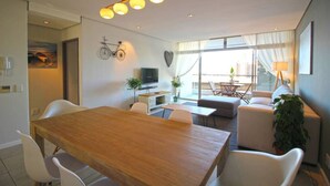 A modern living room with a wooden dining table, white chairs, a sofa, and a television. There is a bicycle decoration on the wall and a plant on the floor. - Upmarket 2bed City Retreat Close to V A Waterfront (Cape Town)