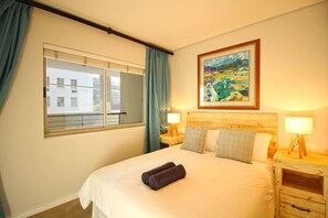 A hotel room with a large bed, two bedside lamps, a painting on the wall, a window with curtains, and a view of buildings outside. - Upmarket 2bed City Retreat Close to V A Waterfront (Cape Town)