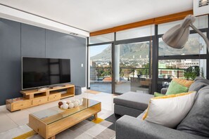 Een moderne woonkamer met een grote flatscreen-televisie, een glazen salontafel en een grijze bank met kleurrijke kussens.