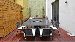 Una terraza de madera con una mesa y sillas, rodeada por un muro con textura y una puerta.