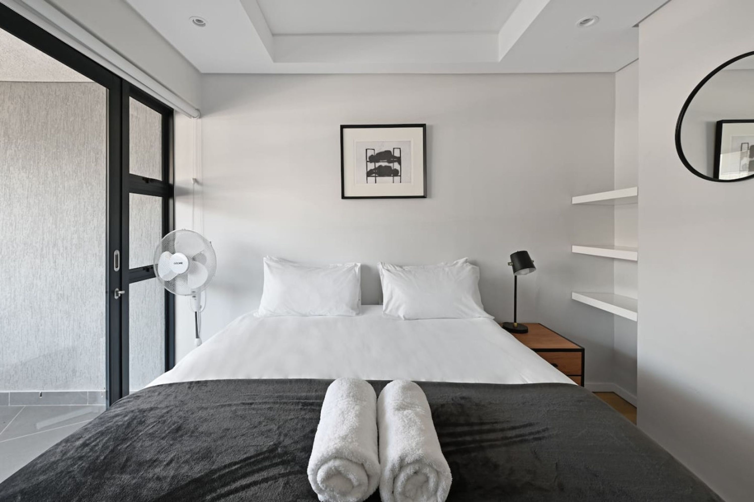 Ein modernes Hotelzimmer mit einem großen Bett, weißen Leinentextilien, einem Ventilator, einer Nachttischlampe und einem gerahmten Bild an der Wand.
