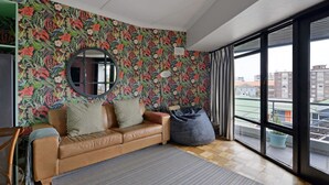 A modern living room with a floral wallpaper, a leather sofa, a round mirror, and a large window offering a view of buildings. - Charming 2-bed Central Green Point Stadium (Cape Town)