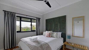 A bedroom with a bed, a ceiling fan, a window with curtains, a bedside table with a lamp, and a wooden bench. - Charming 2-bed Central Green Point Stadium (Cape Town)