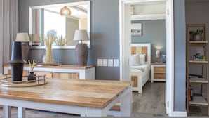 Een moderne woonkamer met een houten salontafel, twee lampen en een grote spiegel die de ruimte weerspiegelt. Een deur leidt naar een slaapkamer met een bed en een nachtkastje.