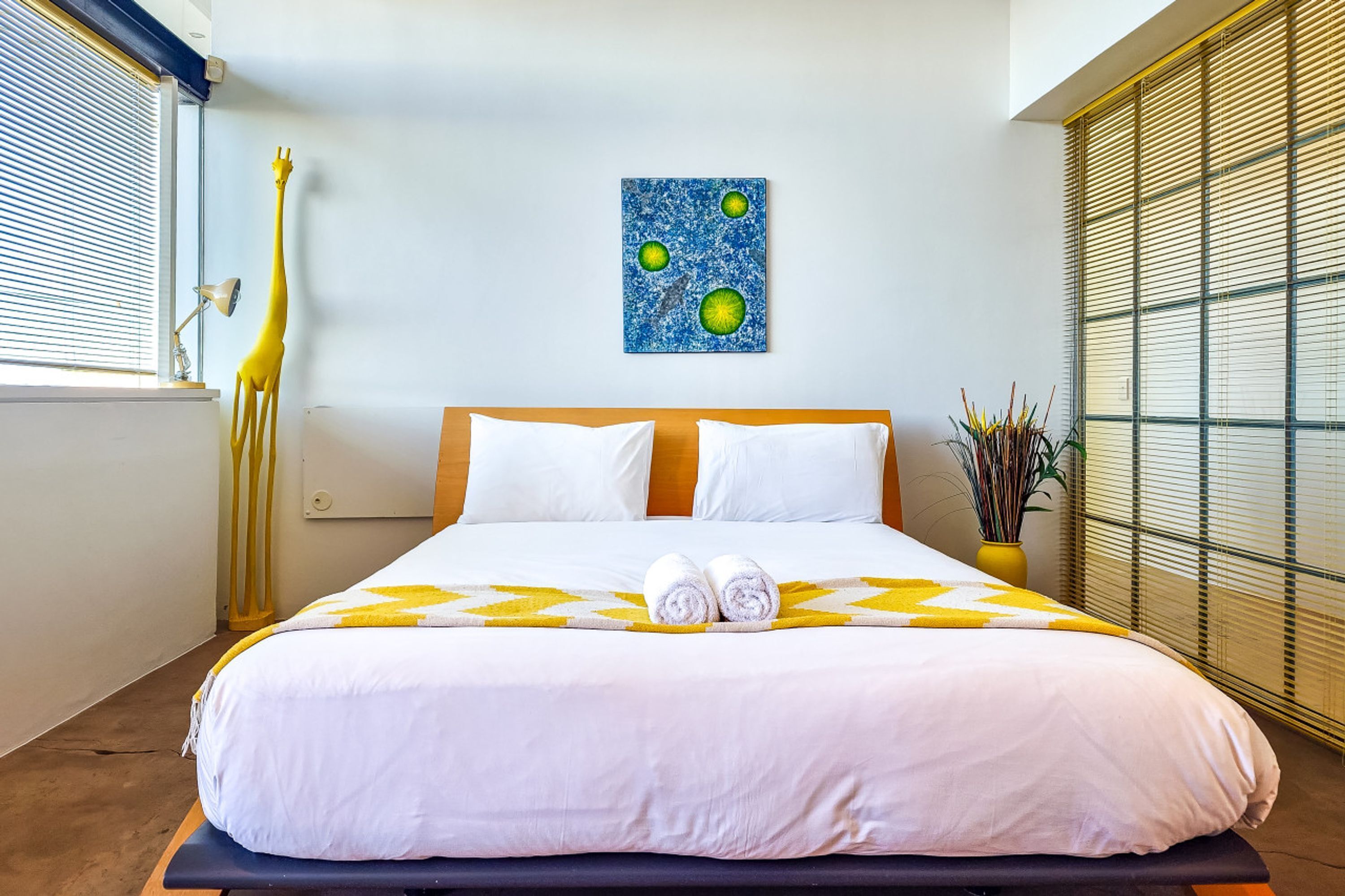 Una cama bien tendida con ropa de cama blanca y amarilla, una escultura de jirafa, una obra de arte en la pared y una ventana con persianas.