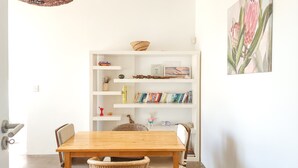 A dining area with a wooden table and wicker chairs, a white bookshelf with various items, and a painting of flowers on the wall. - Green Point Peaceful Retreat Off-grid (Cape Town)