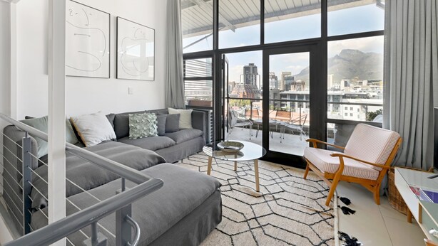 Ein modernes Wohnzimmer mit grauem Sofa, einem kleinen runden Tisch und einem Korbstuhl. Ein großes Fenster bietet Blick auf die Stadt.