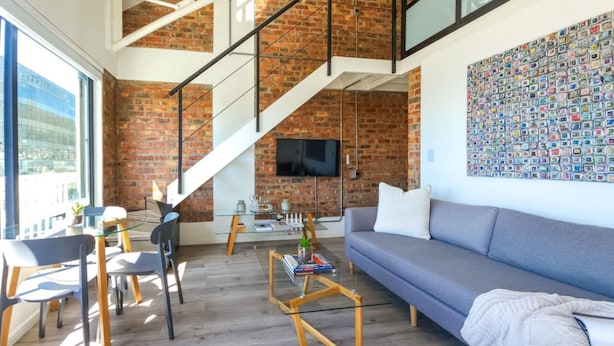 Un salon moderne avec un mur en briques, un escalier, un téléviseur à écran plat, un canapé gris et une table basse en verre.
