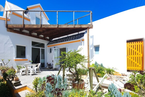 Un bâtiment blanc doté d’une terrasse en bois, d’un patio couvert et de diverses plantes en pot.