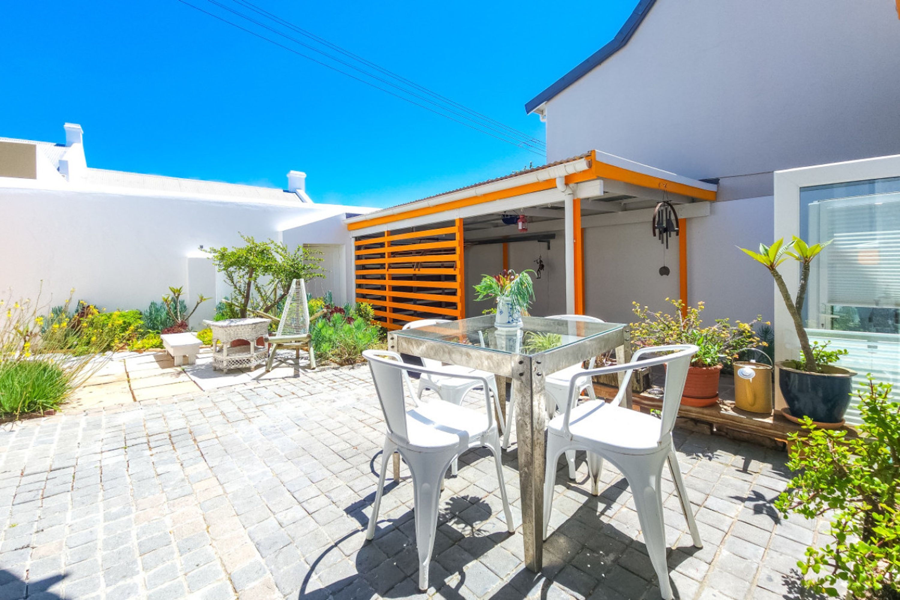 A patio with a glass table and white chairs, surrounded by potted plants and a white fence.