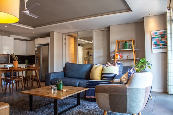 Un salon moderne avec un canapé, une table basse et des fauteuils. On y trouve également un coin cuisine avec un réfrigérateur et des placards. Un ventilateur de plafond est visible, et une plante est posée sur la table basse.