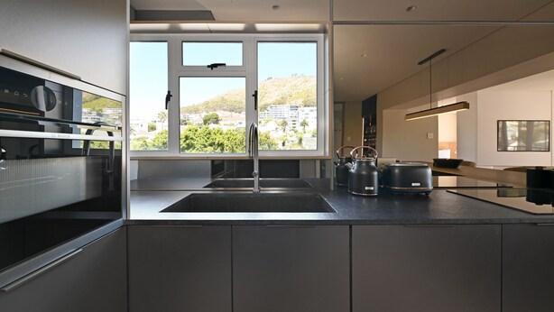 A modern kitchen with a large window, stainless steel appliances, and a minimalist design. - Elegant One Bed Sea Point Mountain Views (Cape Town)